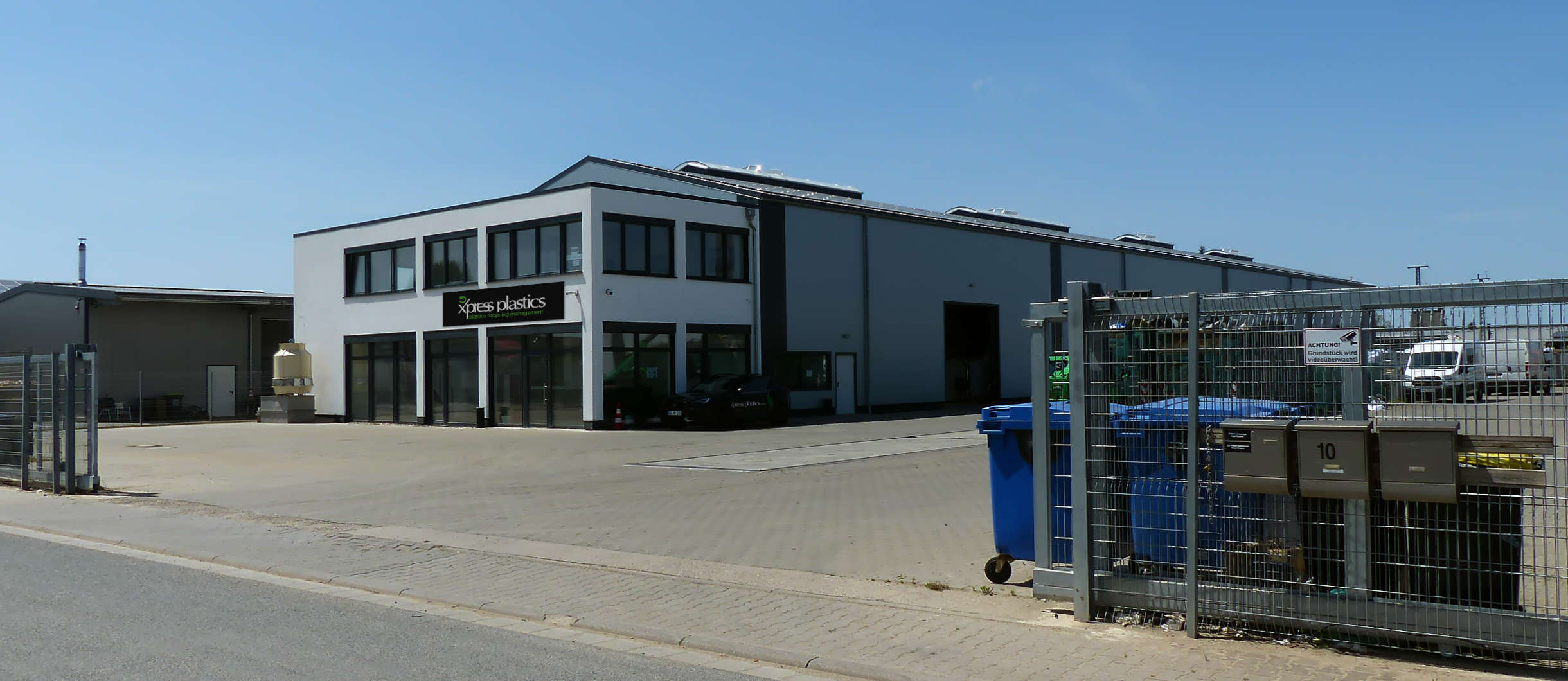Blick auf den Entsorgungsfachbetrieb aus der Einfahrtsrichtung. Weißes Bürogebäude mit dem Firmenlogo Xpress Plastics GmbH über dem Hauseingang. Weiter hinten steht die Produktionshalle angeschlossen. Sie dient zur Verarbeitung von Kunststoff und Plastik.