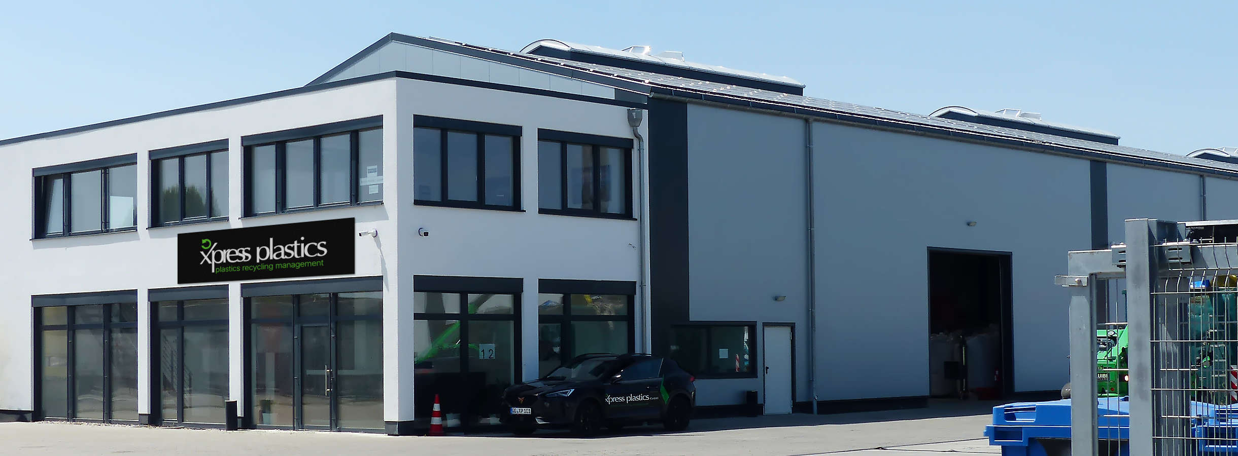 Aus der Zufahrt ist das weiße Bürogebäude des Entsorgungsfachbetriebs Xpress Plastics GmbH für Plastik und Kunststoff zu sehen. Es grenzt an eine große hellgraue Produktionshalle für Rohstoff-Recycling, Lagerung und Handel. Firmenauto parkt daneben.