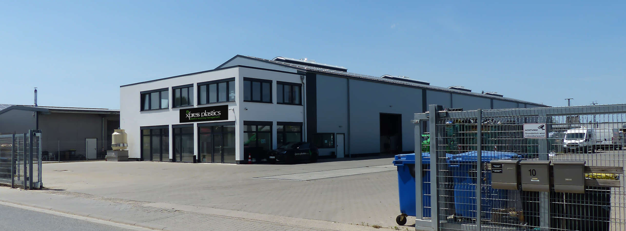 Einfahrtstor des Entsorgungsfachbetriebs Xpress Plastics GmbH ist offen. Am Zaun stehen Abfallcontainer aus Kunststoff. PKW parkt neben einem weißen Bürogebäude, dem folgt eine große hellgraue Produktionshalle für Plastik-Recycling, -Lagerung, -Handel.