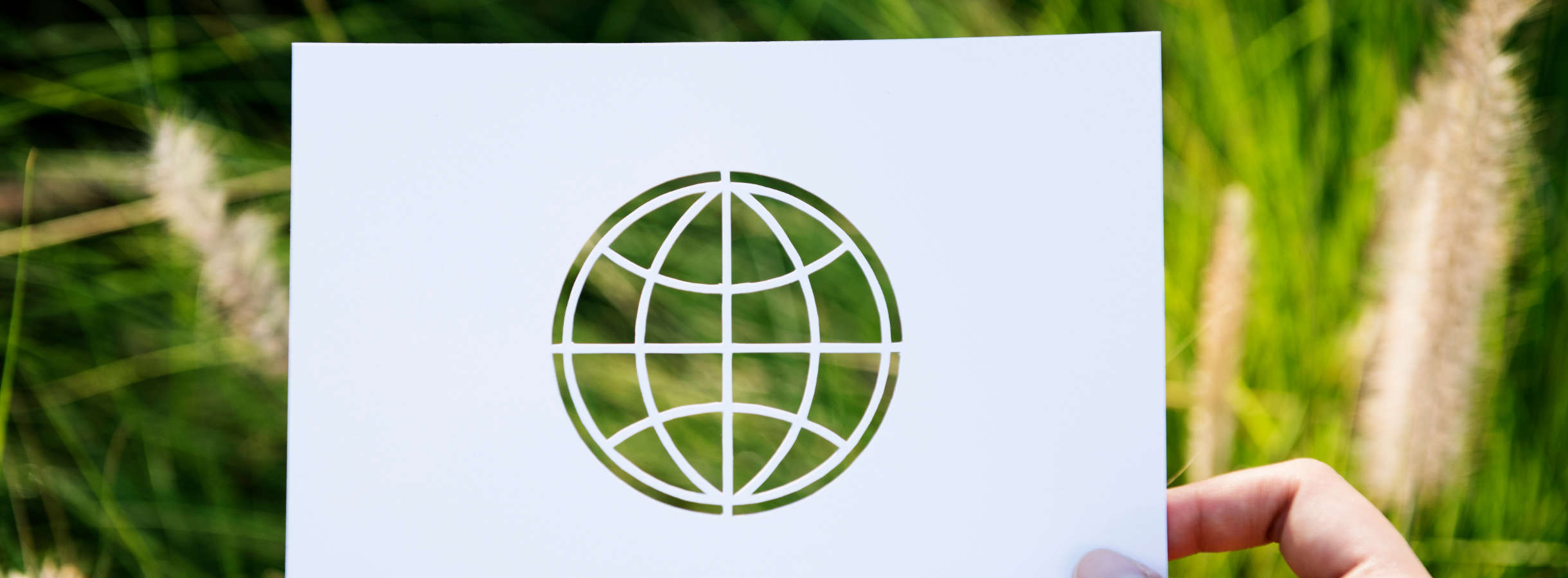 Hohes saftiges grünes Gras füllt den Hintergrund der Abbildung für Umweltschutz aus. Im Vordergrund halten die Finger einer Hand einen recycelten weißen Kunststoff. In der Mitte ist das runde Symbol des Planeten Erde ausgeschnitten.