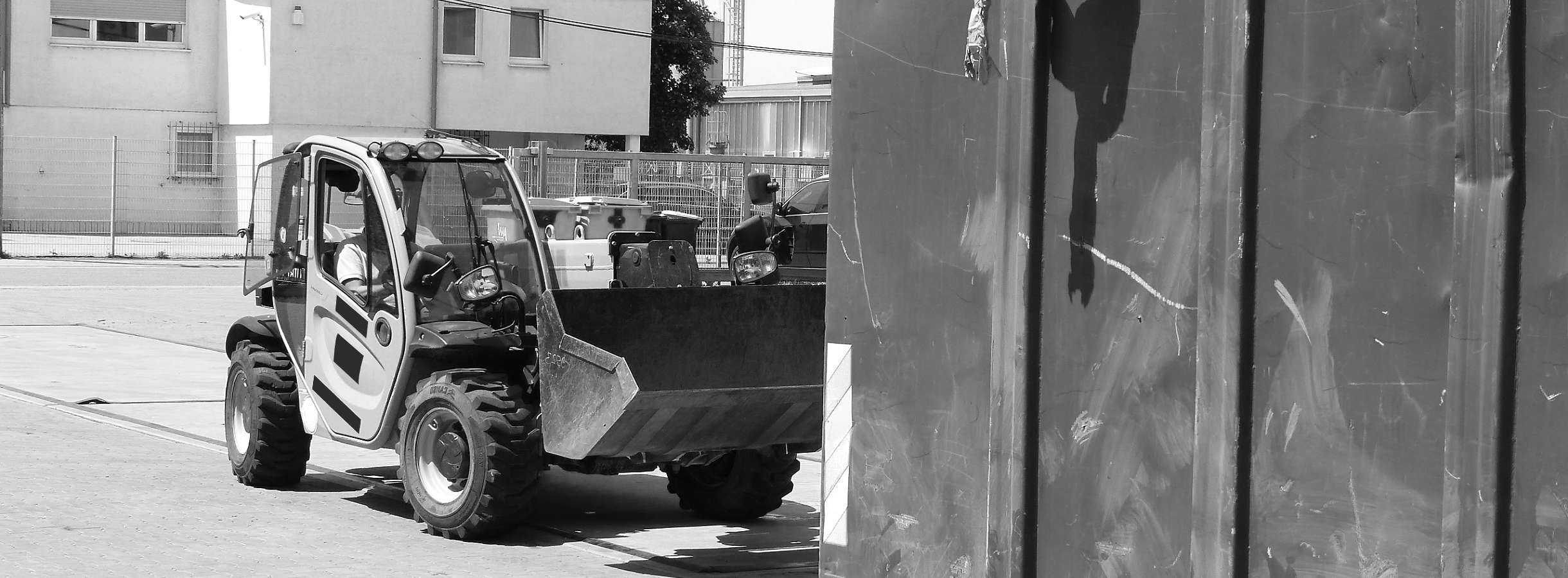 Mitarbeiter bedient einen mit Plastikabfall gefüllten Radlader. Er hat Kunstoffreste zum Recyceln in einen Container abgeladen. Kunststofffetzen hängen aus dem Container für Plastikrecycling herunter. Radlader fährt eine Kunstsoffabfallladung holen.