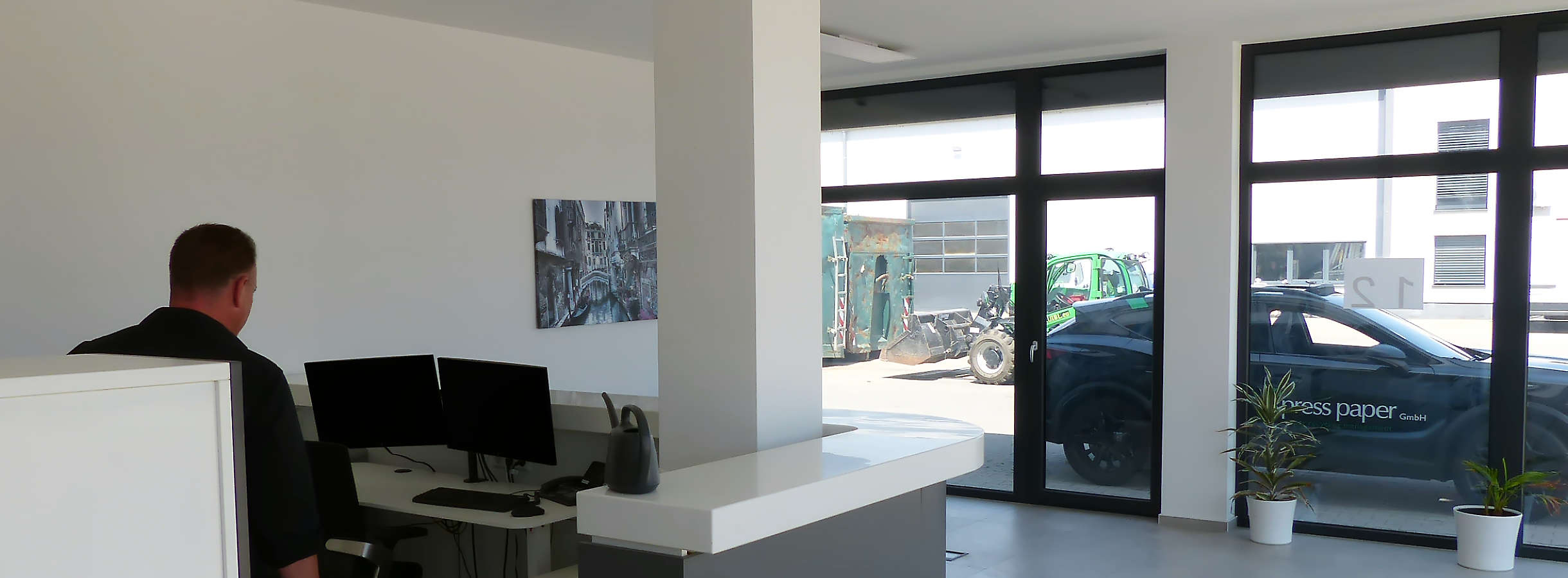 Empfangsbereich des Recyclingunternehmens Xpress Plastics GmbH mit weiten offenen Vitrinen. Durch die großen Fensterscheiben ist der Seitenhof der Firma für Plastik- und Kunststoffentsorgung zu sehen. Dort steht ein grüner Radlader neben einem Container.