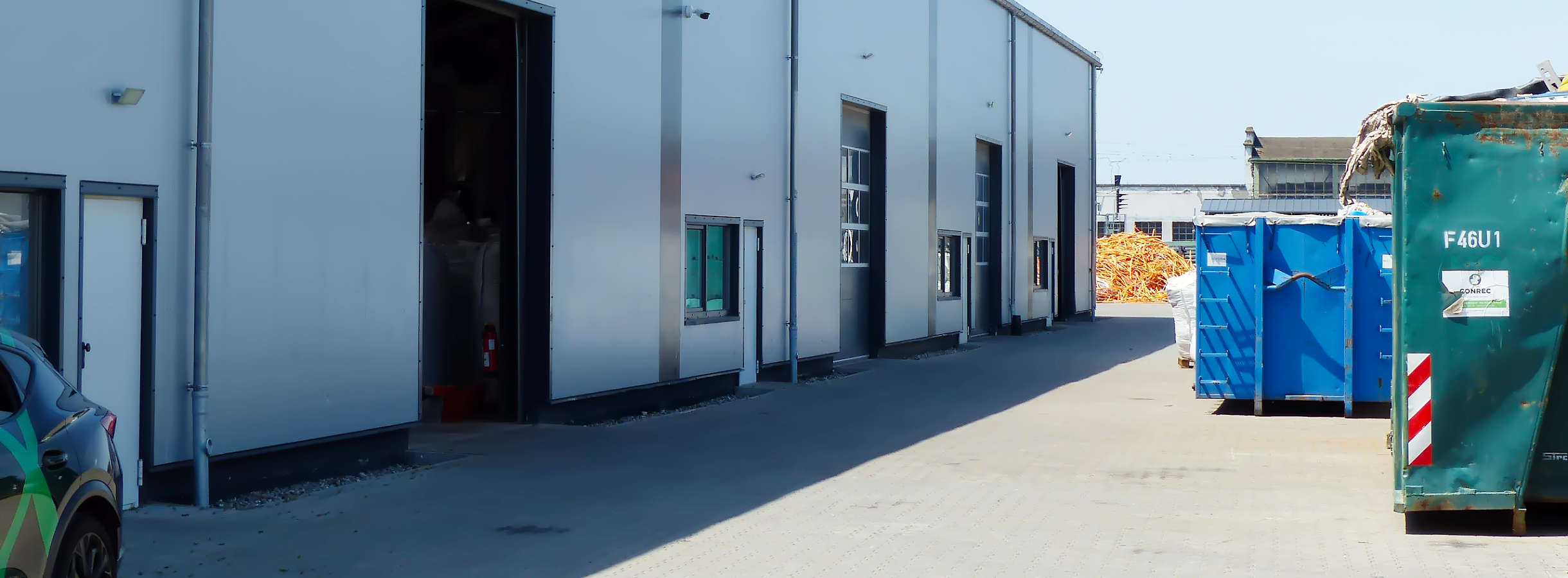 Graue Produktionshalle der Xpress Plastics GmbH für Plastik-Recycling. Am Ende des Geländes befindet sich die Ablagefläche für sortierten Kunststoff- und Plastikabfall. Gegenüber dem Gebäude stehen große Container mit abgeholten unsortierten Plastik.