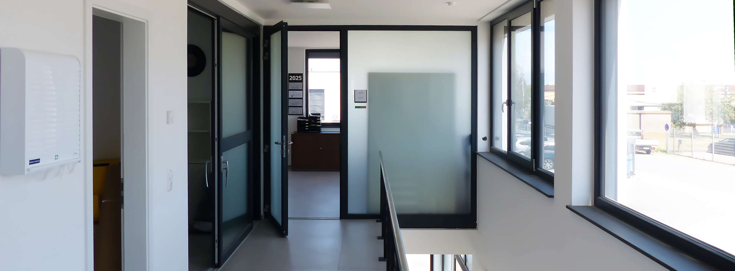 Ein langer heller Flur verbindet mehrere Büroräume vom Entsorgungs- und Recyclingunternehmen Xpress Plastics GmbH. Türen mit Milchglas lassen Licht durch und sorgen für Privatsphäre. Rechts unter den großen Fenstern führt eine Treppe zum Ausgang.
