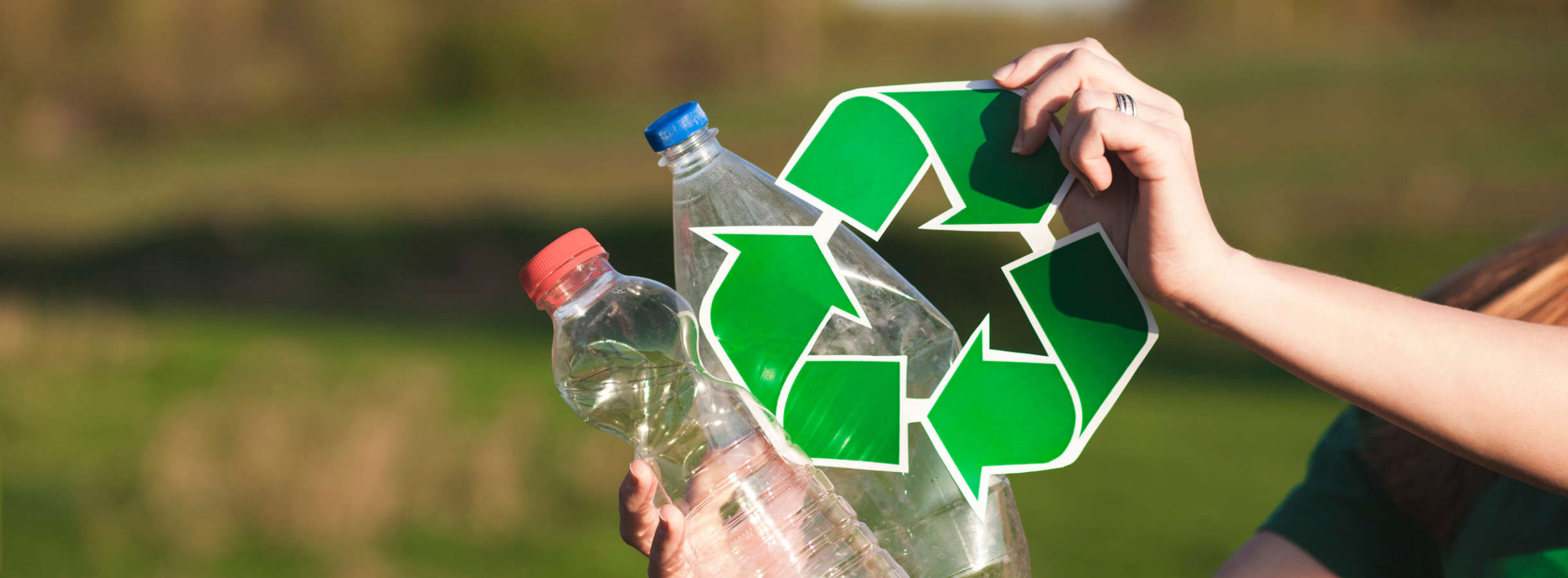 Umweltschützerin sammelt Kunststoffabfall und Plastikmüll in der Hamburger Natur. Sie hält zwei Plastikflaschen in der einen Hand, in der zweiten Hand das Recyclingsymbol mit drei grünen Pfeilen im Kreis angeordnet, die für den Recyclingkreislauf stehen.