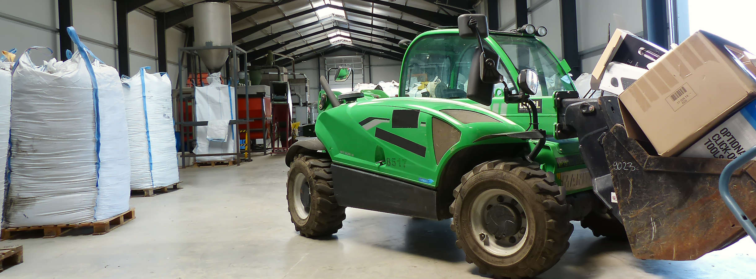 Grüner Radlader mit Verpackungen gefüllt steht in der Produktionshalle vom Xpress Plastics Recyclingunternehmen. In der Halle werden Kunststoffabfälle und Plastikreste zu hochwertigem Mahlgut verarbeitet. Big Bags werden mit Kunststoff-Granulat gefüllt.